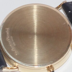 May include: The back of a gold-tone wristwatch with a black leather band. The watch case has the inscription "Patek Philippe Geneve" on the back.