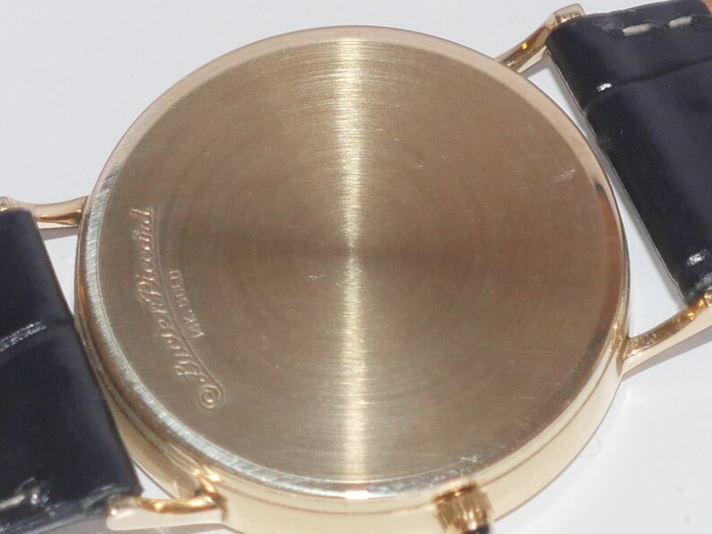 May include: The back of a gold-tone wristwatch with a black leather band. The watch case has the inscription "Patek Philippe Geneve" on the back.