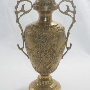 May include: A gold-toned metal vase with ornate detailing and two handles. The vase has a round body and a wide base.