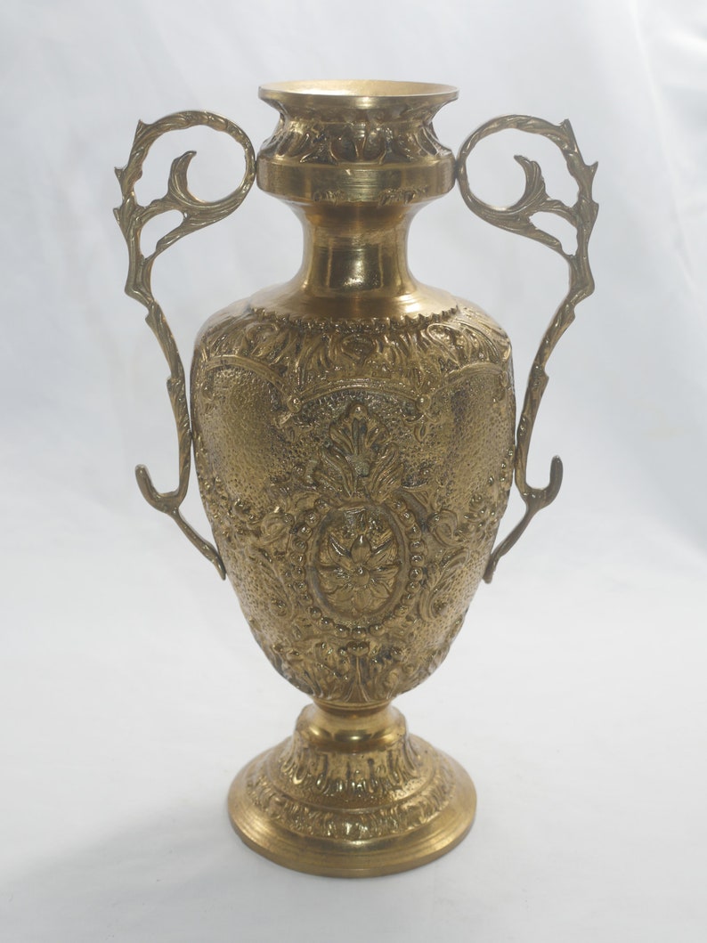 May include: A gold-toned metal vase with ornate detailing and two handles. The vase has a round body and a wide base.