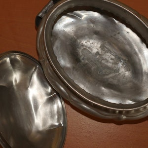 May include: An oval-shaped metallic serving dish with a matching lid placed beside it. The container features a polished, reflective silver-toned surface with a slightly hammered texture and a decorative scalloped rim along the outer edge.