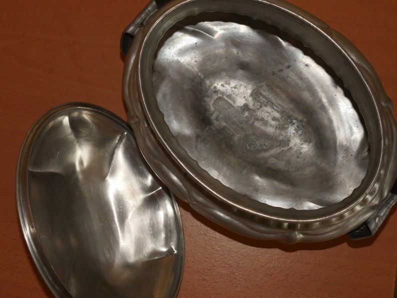 May include: An oval-shaped metallic serving dish with a matching lid placed beside it. The container features a polished, reflective silver-toned surface with a slightly hammered texture and a decorative scalloped rim along the outer edge.