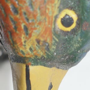 May include: Close-up of a painted duck decoy with a yellow beak and a black eye. The duck's head is green and brown with yellow and orange markings.
