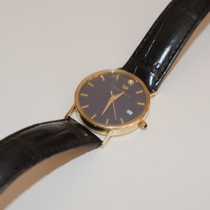 May include: A gold-tone wristwatch with a black leather band and a dark blue face. The watch has a diamond at the 12 o'clock position and the text "LP" on the face.