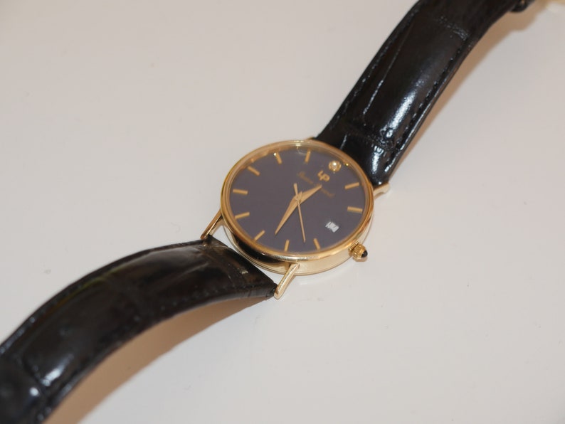 May include: A gold-tone wristwatch with a black leather band and a dark blue face. The watch has a diamond at the 12 o'clock position and the text "LP" on the face.