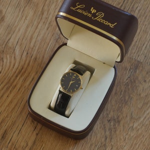 May include: A gold-tone wristwatch with a black face and black leather band in a brown box with the text "LP Lucien Piccard" on the top.