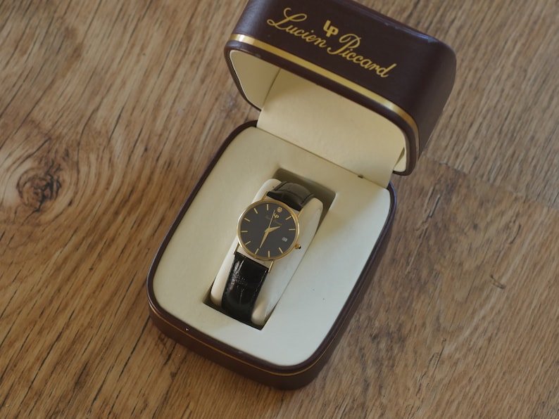 May include: A gold-tone wristwatch with a black face and black leather band in a brown box with the text "LP Lucien Piccard" on the top.