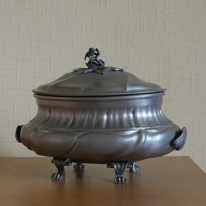 May include: A dark gray metal tureen with a rounded body, a matching lid featuring a small sculpted figure on top, and four ornate feet. The vessel has a wide, curved rim and two side handles, showcasing a smooth, matte metallic finish throughout.