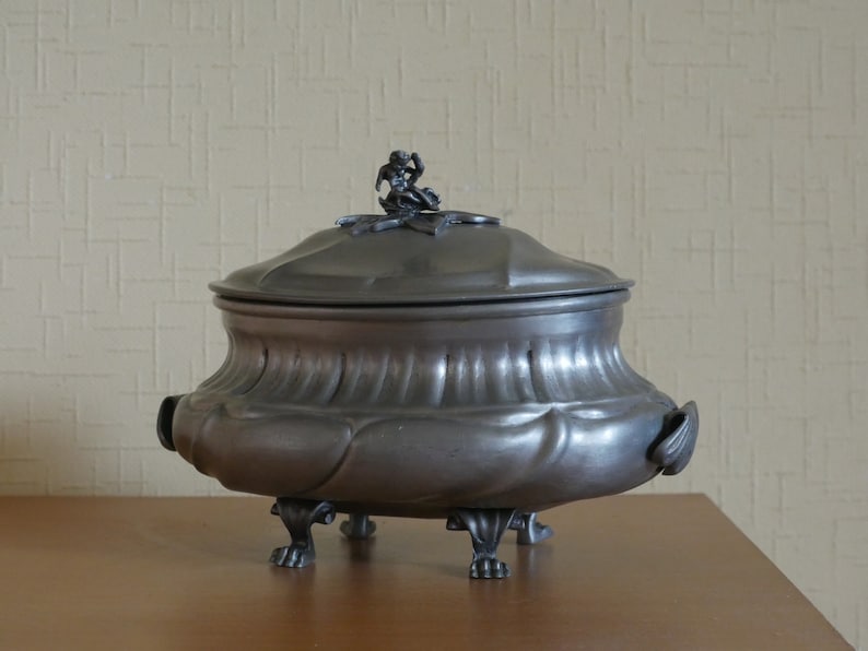 May include: A dark gray metal tureen with a rounded body, a matching lid featuring a small sculpted figure on top, and four ornate feet. The vessel has a wide, curved rim and two side handles, showcasing a smooth, matte metallic finish throughout.