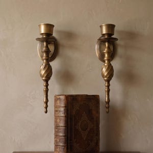 Könnte beinhalten: Zwei passende Wandleuchter aus Messing mit einem konisch zulaufenden, gedrehten Stiel und einem runden Kerzenhalter. Unter den Leuchtern steht ein dickes, ledergebundenes antikes Buch mit dem Titel Chronicles of Empires auf einem dunklen Holztisch.