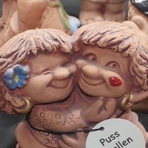Pu&ograve; includere: Due figurine in ceramica di persone con capelli castani e volti sorridenti. Le figurine si abbracciano. La figurina a sinistra ha un fiore blu nei capelli. La figurina a destra ha il rossetto rosso. Le figurine sono etichettate "Puss" e "Allen".