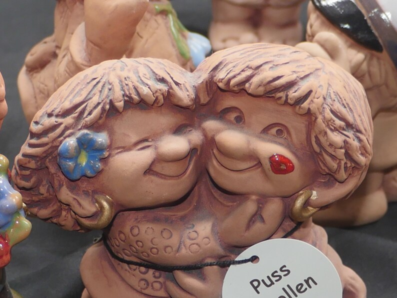 Pu&ograve; includere: Due figurine in ceramica di persone con capelli castani e volti sorridenti. Le figurine si abbracciano. La figurina a sinistra ha un fiore blu nei capelli. La figurina a destra ha il rossetto rosso. Le figurine sono etichettate "Puss" e "Allen".