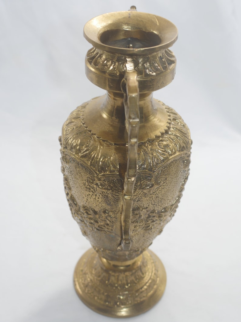 May include: A gold metal vase with an ornate design. The vase has a wide mouth and a narrow base. It is decorated with intricate floral patterns.