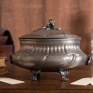 May include: A metallic gray lidded tureen with a rounded body, featuring a sculpted finial on the lid and four ornate feet. The vessel has a smooth, curved design with integrated side handles, presenting a polished, uniform finish throughout.