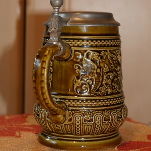 Może przedstawiać: Brązowy ceramiczny kufel do piwa z pokrywką z cyny i srebrnym uchwytem. Kufel jest ozdobiony kwiatowymi i zwiniętymi wzorami.