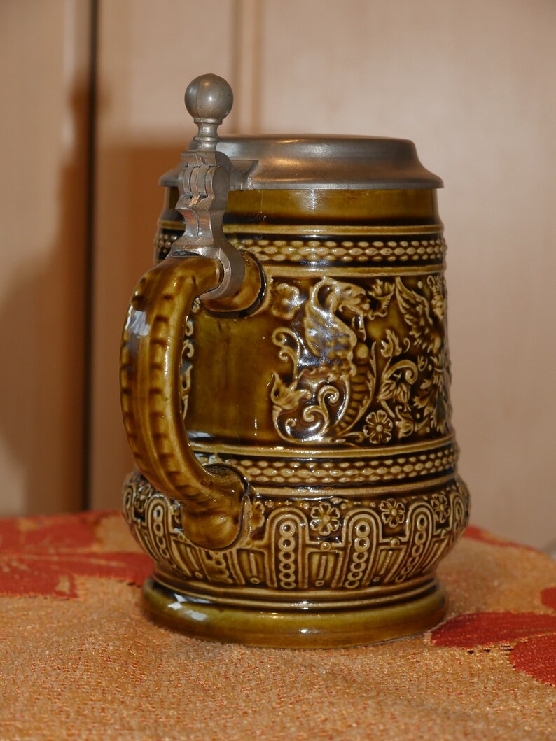 Może przedstawiać: Brązowy ceramiczny kufel do piwa z pokrywką z cyny i srebrnym uchwytem. Kufel jest ozdobiony kwiatowymi i zwiniętymi wzorami.
