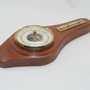 May include: A wooden barometer with a brass face and a thermometer on the side. The barometer face has a white background with black markings and the word "RECORDER" printed on it.