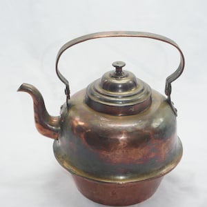 Op de afbeelding: Een vintage koperen waterkoker met een verweerde afwerking en een rond lichaam. De waterkoker heeft een enkele handgreep en een deksel met een knop.