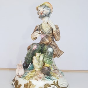 Pode incluir: Uma estatuinha de porcelana de um homem com barba e chapéu, sentado em uma base com um design floral. Ele está segurando um pequeno pássaro na mão e tem um pequeno porco aos seus pés.