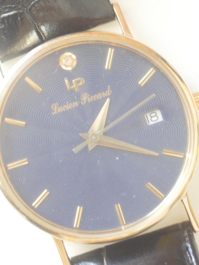 May include: A gold-tone wristwatch with a blue face and gold-tone hands. The watch has a diamond at the 12 o'clock position and the text 'Lucien Piccard' on the face.