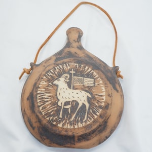 Op de afbeelding: Een bruine keramische fles met een leren riem. De fles heeft een ontwerp met een ram met een kruis en een vlag op zijn rug.