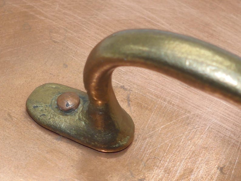 May include: Close-up of a copper pot with a brass handle. The handle is curved and has a small, round knob on the top.