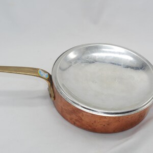 May include: A copper saucepan with a brass handle and a silver-colored aluminum insert. The saucepan has a hammered copper finish.
