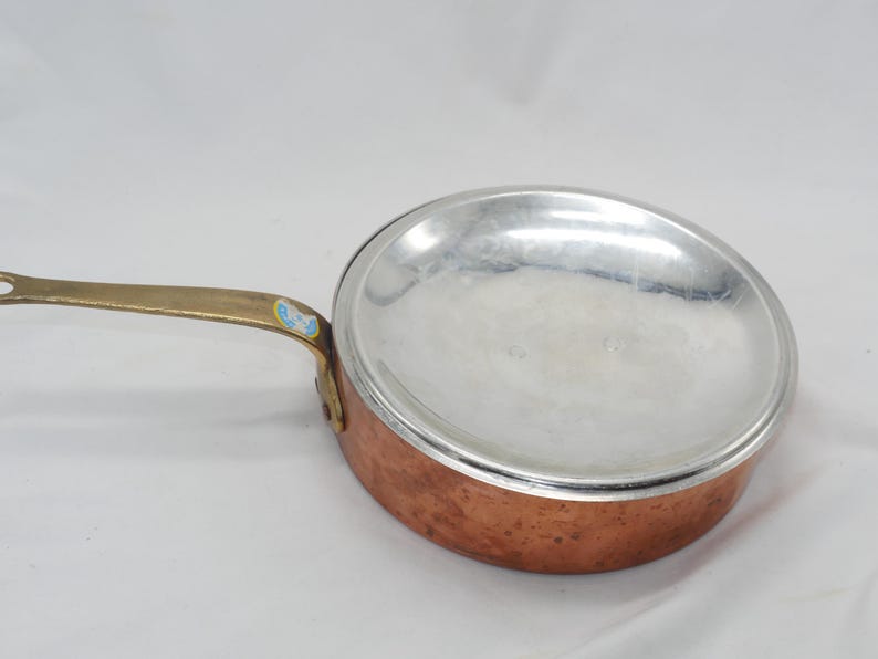 May include: A copper saucepan with a brass handle and a silver-colored aluminum insert. The saucepan has a hammered copper finish.