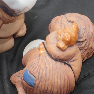 Pu&ograve; includere: Una statuetta in ceramica di un bambino che dorme con un cappello marrone e arancione. Il bambino &egrave; sdraiato di lato con le braccia avvolte attorno a un piccolo oggetto marrone. La statuetta &egrave; dipinta in tonalit&agrave; di marrone, arancione e blu.