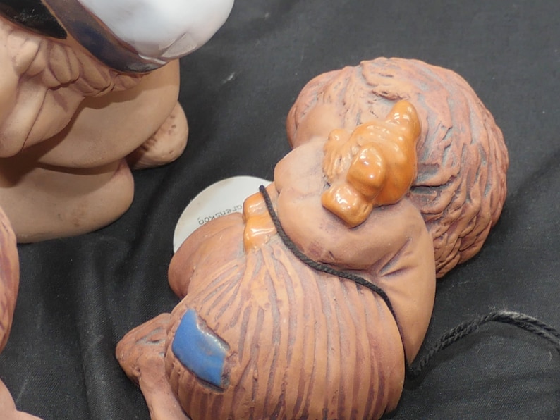 Pu&ograve; includere: Una statuetta in ceramica di un bambino che dorme con un cappello marrone e arancione. Il bambino &egrave; sdraiato di lato con le braccia avvolte attorno a un piccolo oggetto marrone. La statuetta &egrave; dipinta in tonalit&agrave; di marrone, arancione e blu.