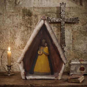 May include: A wooden shrine featuring a carved figure wearing a yellow robe and a dark cloak, housed within a triangular, gabled frame with a weathered, light-colored finish. The figure stands centrally inside the open, dark-toned interior.