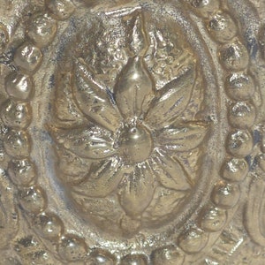 May include: Close-up of a gold-toned metal ornament with a detailed floral design. The center of the ornament features a raised flower with a circular center.