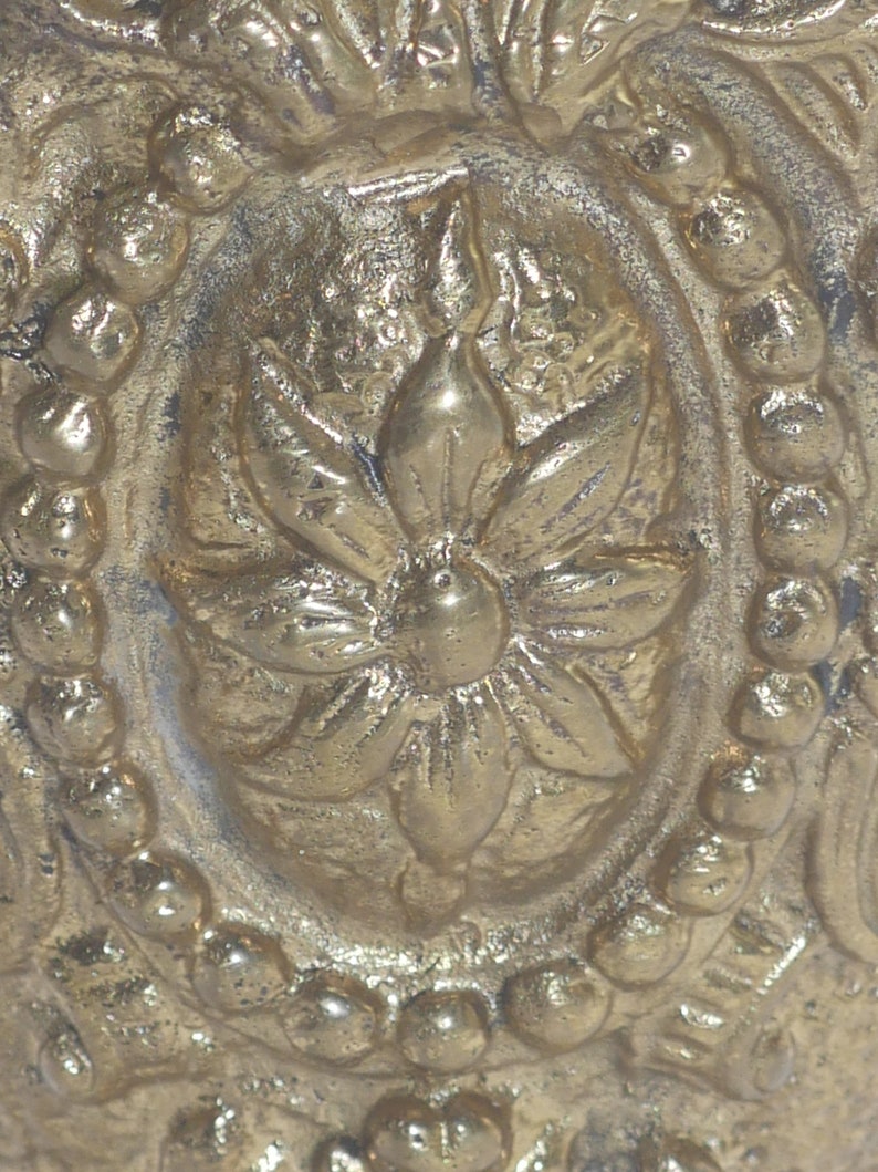 May include: Close-up of a gold-toned metal ornament with a detailed floral design. The center of the ornament features a raised flower with a circular center.