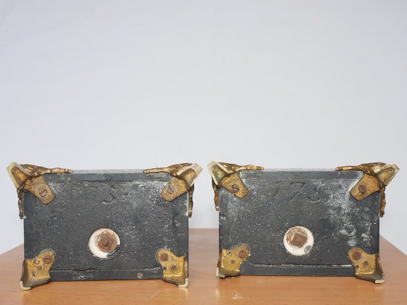 May include: Two black bookends with gold accents and a white marble base. The bookends are shaped like a pair of hands holding a book. The bookends have the numbers 1773 and 1773 inscribed on them.