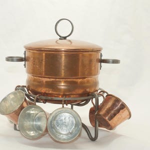 Può includere: Una pentola per fonduta in rame con coperchio e piccole tazze. La pentola ha un manico sul coperchio e due manici laterali. Le tazze sono sospese a un supporto in ferro battuto.