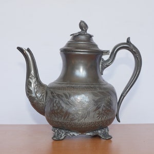 Op de afbeelding: Antieke zilverkleurige theepot met ingewikkelde bloemengravures. De theepot heeft een gebogen tuit, een decoratieve handgreep en een deksel met een kleine knop. Hij staat op drie kleine voetjes en toont een vintage ontwerp.