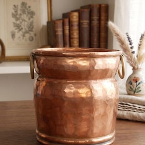 May include: A cylindrical copper bucket featuring a hammered texture throughout its exterior surface. The vessel includes a reinforced rim and two circular metal ring handles attached to opposite sides near the top edge of the container.