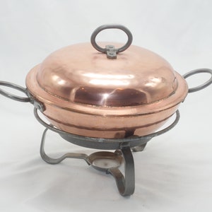 May include: A copper chafing dish with a lid and two handles. The dish sits on a black metal stand with three legs.
