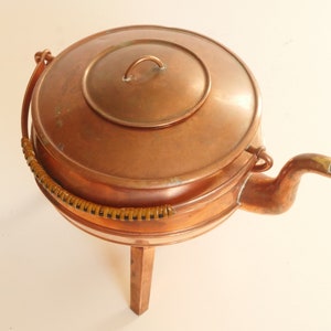 May include: A copper teapot with a lid and a handle, sitting on a wooden stand with three legs. The teapot has a spout and a woven handle.
