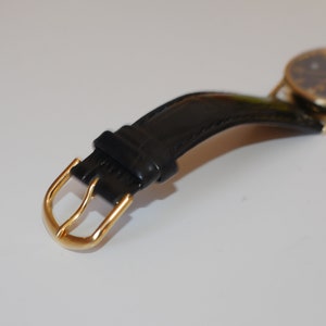 May include: A gold-toned watch with a black leather strap and a gold buckle. The watch face is black with gold hands and markers.