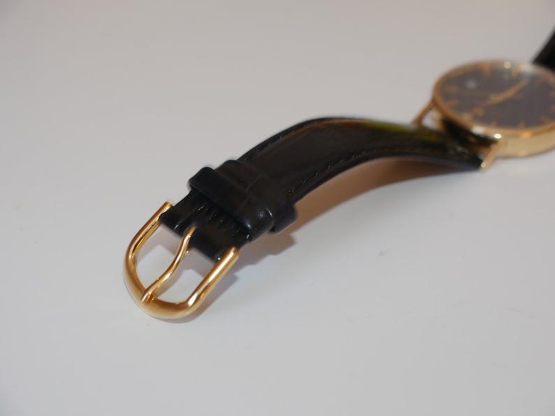 May include: A gold-toned watch with a black leather strap and a gold buckle. The watch face is black with gold hands and markers.