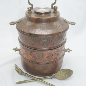 K&ouml;nnte beinhalten: Ein dreist&ouml;ckiger Tiffin-Beh&auml;lter aus Kupfer mit einem verzierten, gravierten Design. Der Beh&auml;lter hat einen Messinggriff und zwei Messingverschl&uuml;sse. Er wird mit einem L&ouml;ffel und einer Gabel aus Messing geliefert.