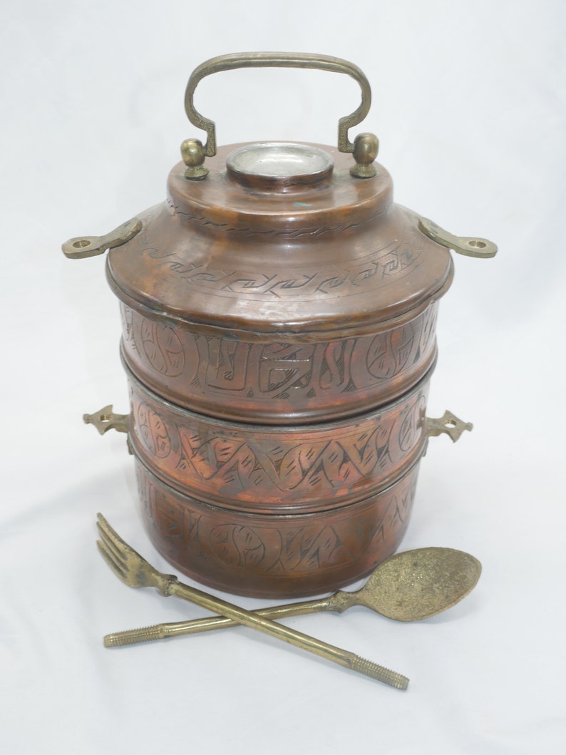 K&ouml;nnte beinhalten: Ein dreist&ouml;ckiger Tiffin-Beh&auml;lter aus Kupfer mit einem verzierten, gravierten Design. Der Beh&auml;lter hat einen Messinggriff und zwei Messingverschl&uuml;sse. Er wird mit einem L&ouml;ffel und einer Gabel aus Messing geliefert.