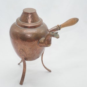 Pode incluir: Uma chaleira de cobre martelado com uma pega de madeira e três pernas. A chaleira tem uma tampa e um bico.