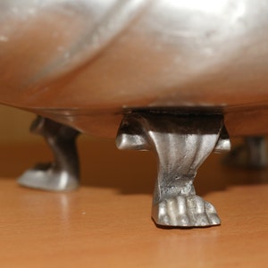 May include: A silver-toned metal bowl featuring three distinct feet shaped like animal paws with visible claws. The bowl has a smooth, reflective surface and a curved base that connects to the detailed, textured feet at the bottom of the object.