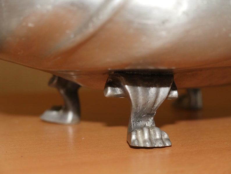 May include: A silver-toned metal bowl featuring three distinct feet shaped like animal paws with visible claws. The bowl has a smooth, reflective surface and a curved base that connects to the detailed, textured feet at the bottom of the object.
