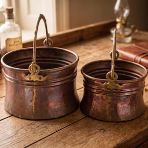 Op de afbeelding: Twee ronde koperen emmers met messing zwenkhandvatten en decoratieve bevestigingen. De emmers hebben een donker, verweerd patina met zichtbare verticale naden en horizontale ribbels langs de zijkanten. De metalen oppervlakken vertonen kleurvariaties.