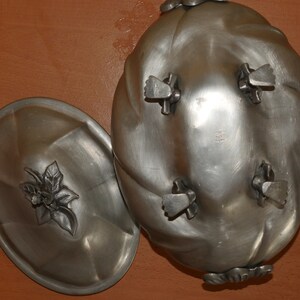 May include: An oval silver-toned metal serving dish with a matching lid. The dish features four feet and a swirled design. The lid is topped with a decorative metal floral finial. The metal has a smooth, matte finish throughout the entire piece.