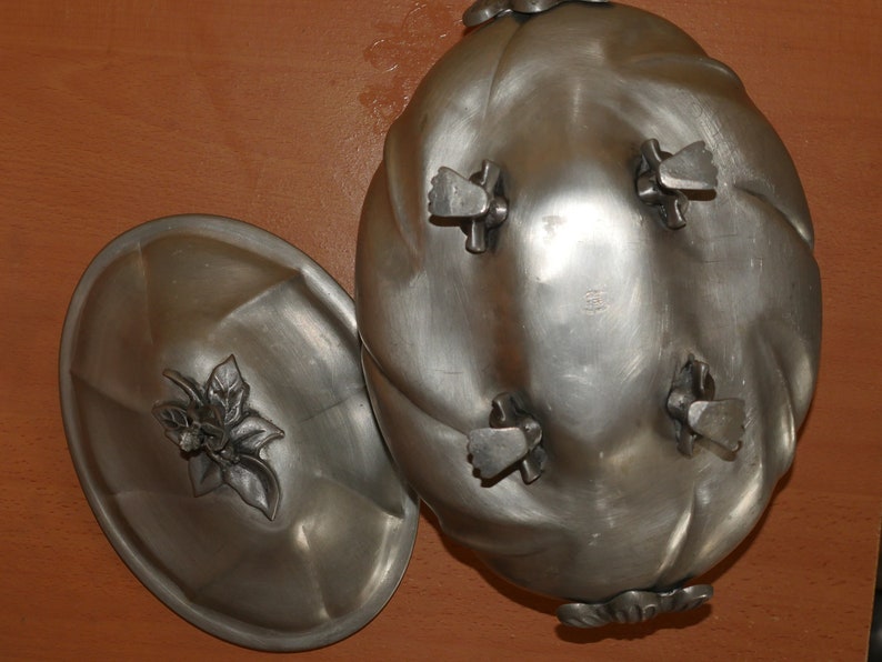 May include: An oval silver-toned metal serving dish with a matching lid. The dish features four feet and a swirled design. The lid is topped with a decorative metal floral finial. The metal has a smooth, matte finish throughout the entire piece.