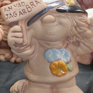 Pu&ograve; includere: Una statuetta in ceramica di un gnomo sorridente che indossa un cappello da marinaio blu e bianco. Lo gnomo tiene un cartello con su scritto "EN VAD JAG &Auml;R BRA!" Lo gnomo indossa una camicia marrone con fiori blu e arancioni.